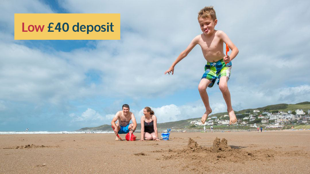 boy jumping on a sandcastle he made whilst holding a spade as his amused parents watch on with Woolacombe hills and Atlantic Ocean in background